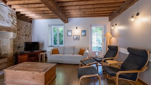 A cozy living room with a stone wall, wooden beams, a white sofa, two armchairs, a wooden coffee table, and a television.