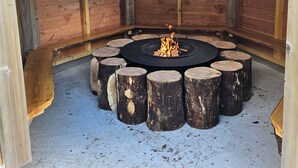 Un hogar circular rodeado de troncos de madera, ubicado en una estructura de madera con techo inclinado y vigas visibles.
