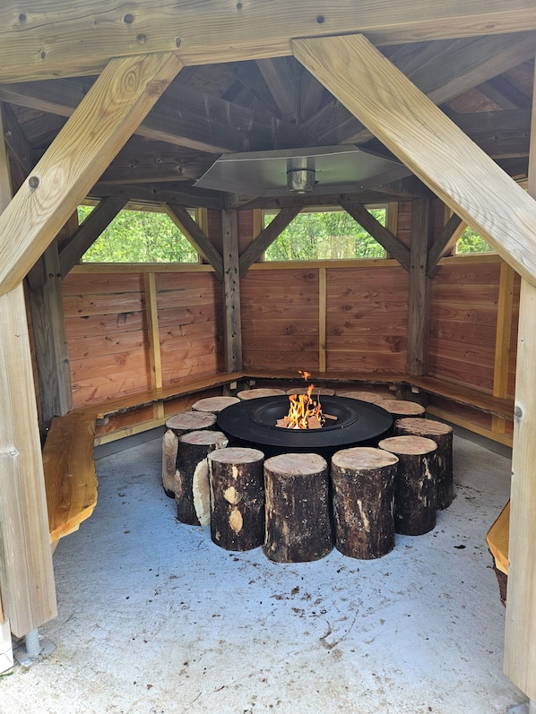 A circular fire pit surrounded by wooden logs, set in a wooden structure with a sloped roof and visible wooden beams.