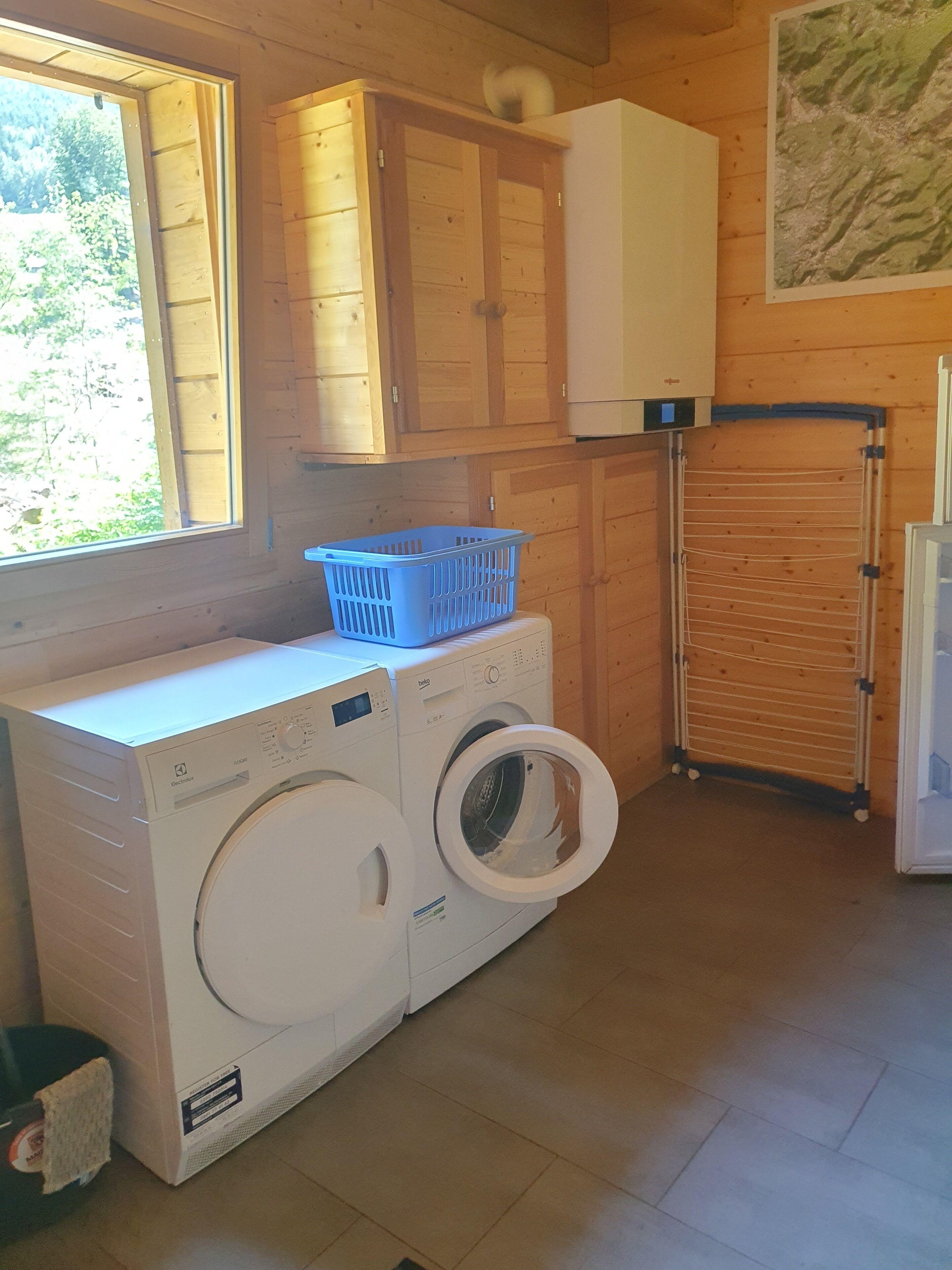 A laundry area with a front-loading washing machine and dryer, wooden cabinets, a drying rack, and a window with a view of greenery.