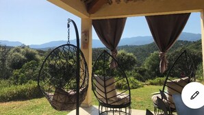 Un espace extérieur aménagé avec une zone de repos couverte, comprenant deux fauteuils suspendus et une table, offrant une vue sur un paysage montagneux.