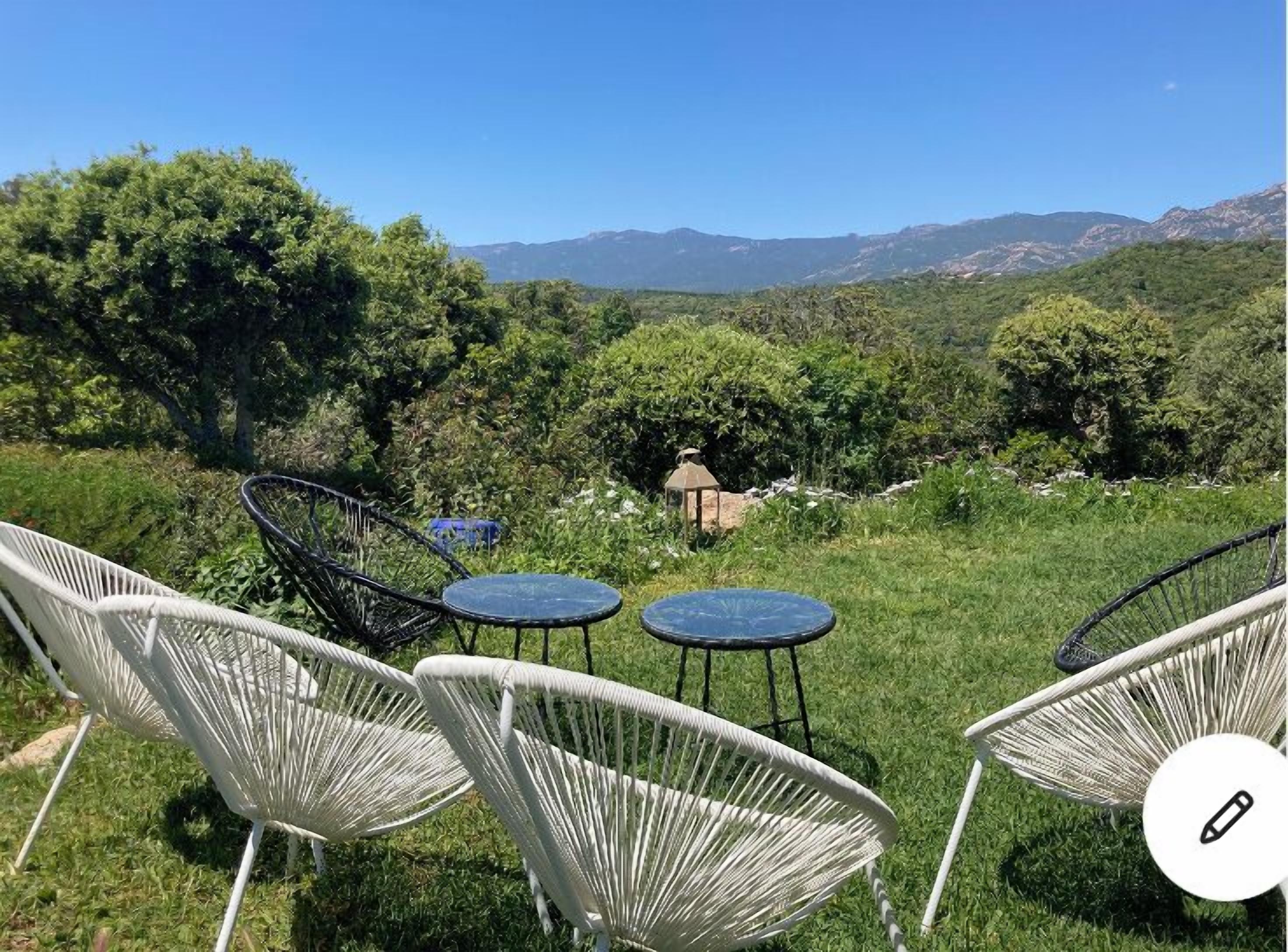 Une terrasse dotée de chaises en osier blanc et de petites tables rondes, offrant une vue sur un paysage verdoyant et luxuriant, avec des arbres et des montagnes à l’horizon.