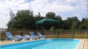 Een rechthoekig zwembad met helderblauw water, omgeven door een houten hek. Er staan meerdere ligstoelen langs de zwembadrand en een groene parasol zorgt voor schaduw.