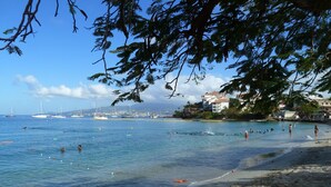 A beach with clear blue water, a sandy shore, and a backdrop of buildings and boats.