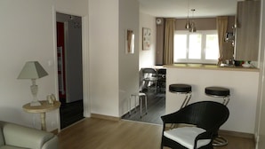 Un salon moderne comprenant un canapé, une table à manger avec des chaises, un coin cuisine avec une fenêtre et un fauteuil en osier noir.