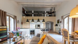 Ein geräumiger Wohnbereich mit einer Couch, einem Glastisch und einem Esstisch mit Stühlen. Es gibt eine Küchenzeile mit modernen Geräten sowie einen Balkon mit Zierelementen.