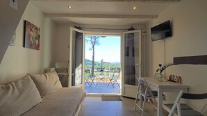 Ein gemütliches Zimmer mit einer Couch, einem Schreibtisch mit Stuhl, einem Fernseher und Blick auf eine Terrasse mit Tisch und Stühlen.
