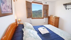 A bedroom with a bed, a wooden dresser, a nightstand with a lamp, and a window with a view of hills. - Villa José 1, 12 Pers, Sea View ! Heated pool,  Wifi,A/C, petfriendly (Comares)