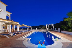 Piscina com espreguiçadeiras e guarda-sóis, cercada por um edifício de paredes brancas e telhado telhado.