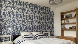 A bedroom with a bed, nightstands, a bedside table, a lamp, a bookshelf, and a wall with a floral pattern.