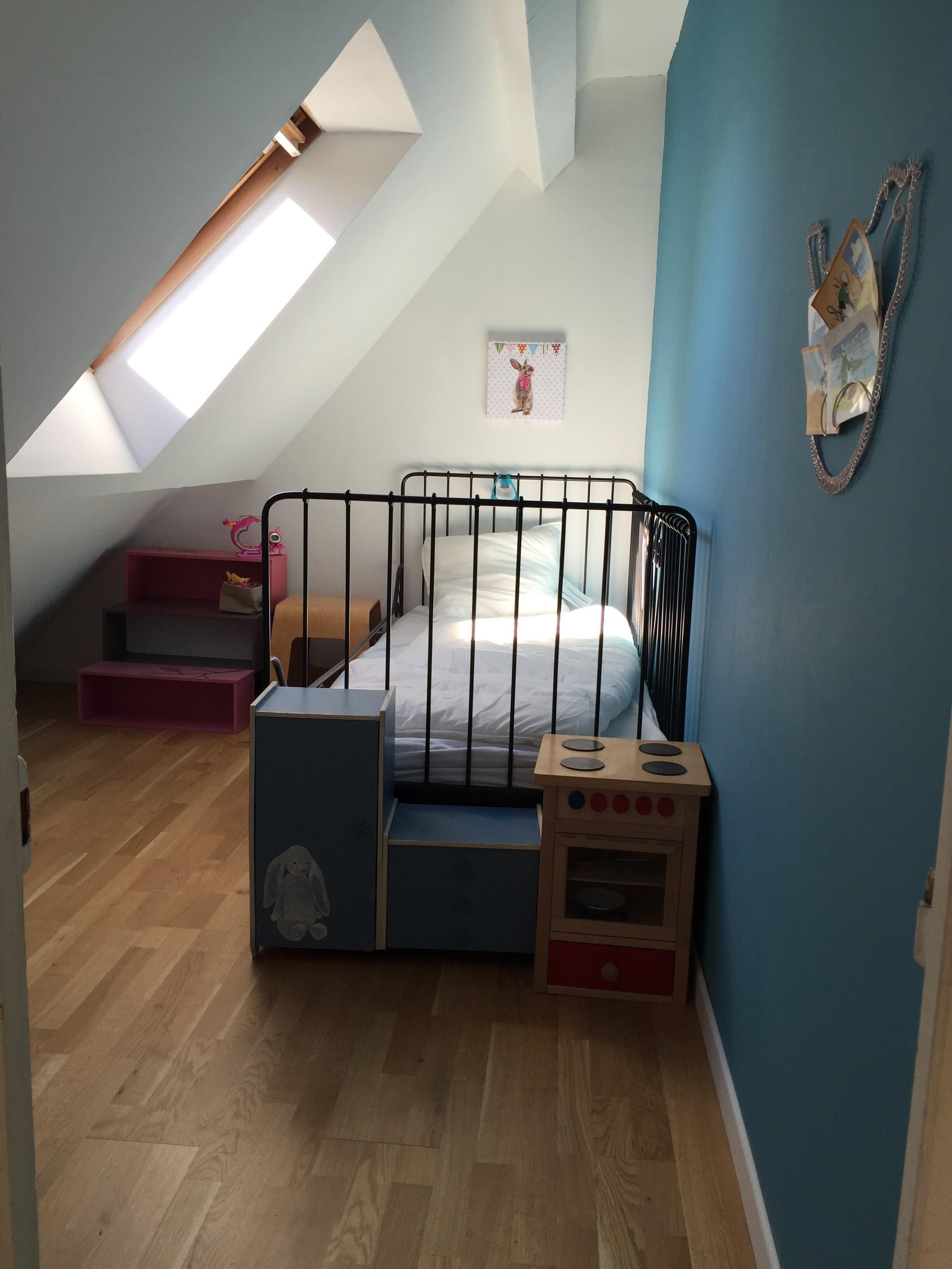 Una camera da letto mansardata con soffitto spiovente, un letto con struttura in metallo, una piccola cucina giocattolo in legno e un velux.