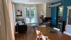 A dining area with a wooden table and chairs, a window with curtains, a radiator, and a small table with a lamp.