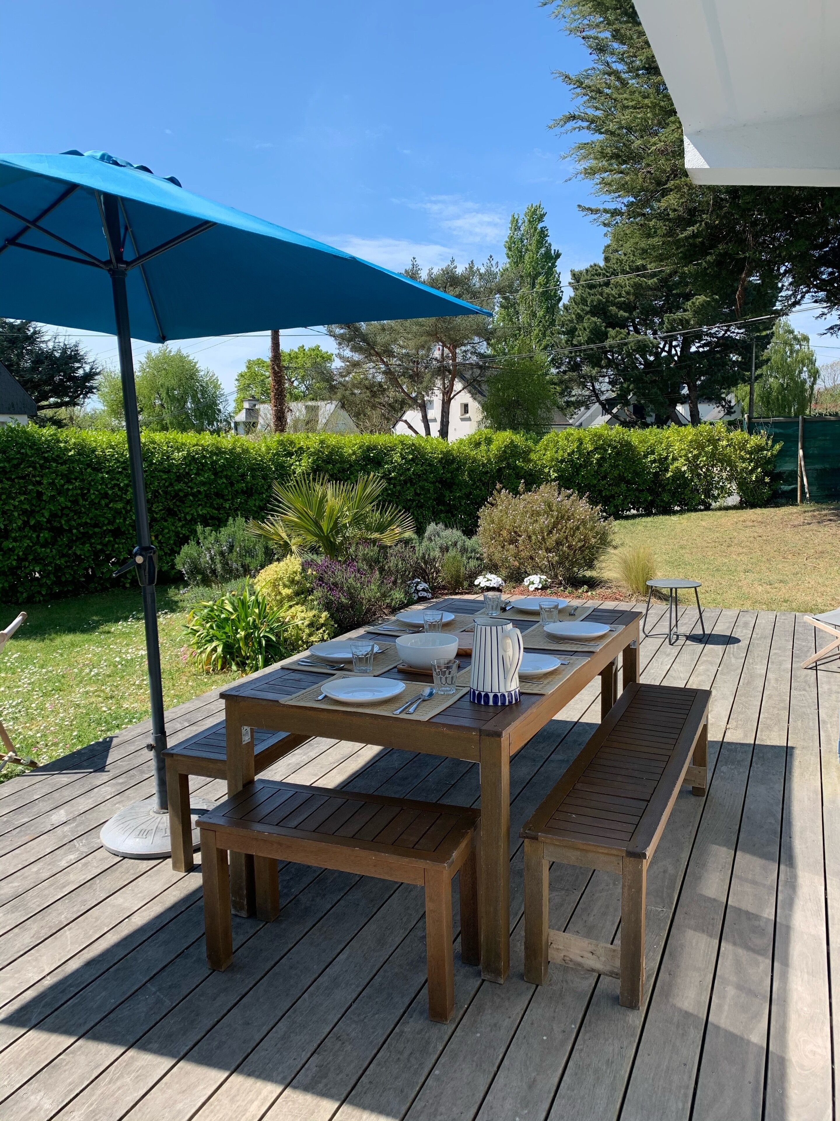 Ein Essbereich im Freien mit Holztischen und -bänken, einem blauen Sonnenschirm und einem gepflegten Garten mit verschiedenen Pflanzen und einer Hecke.