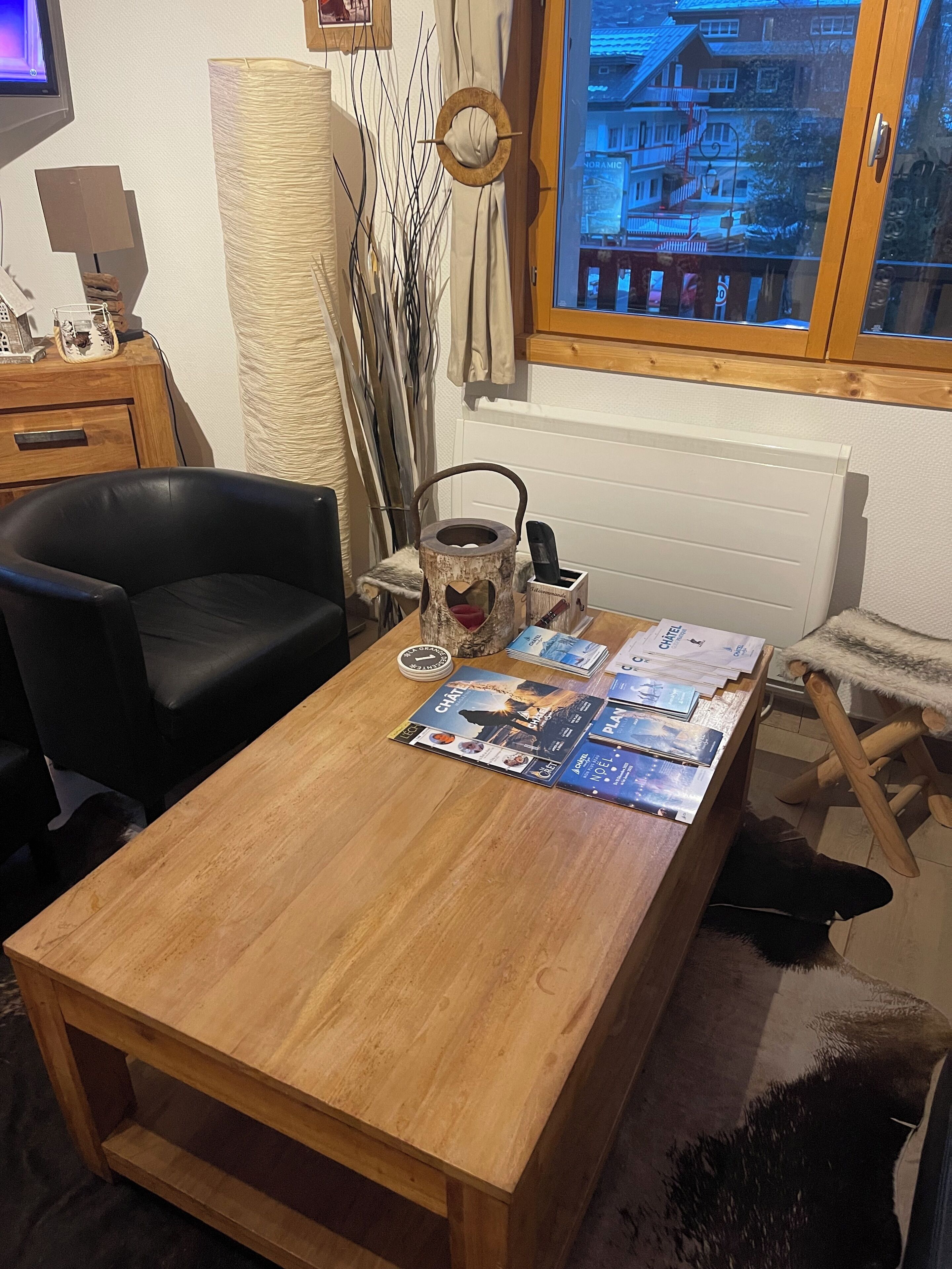 Un tavolo da pranzo in legno con una rivista e un bollitore. Si nota una sedia nera e una poltrona in pelliccia. Da una finestra si vede un edificio all'esterno.