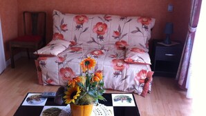 A room with a floral-patterned sofa, a small table with a vase of flowers, a chair, and a nightstand with a lamp.