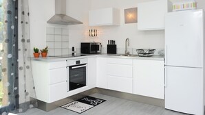 A modern kitchen with white cabinetry, a built-in oven, a microwave, and a refrigerator. There is a window with a curtain, and a small potted plant on the counter. - Apartments in an old renovated sheepfold  (Figari)