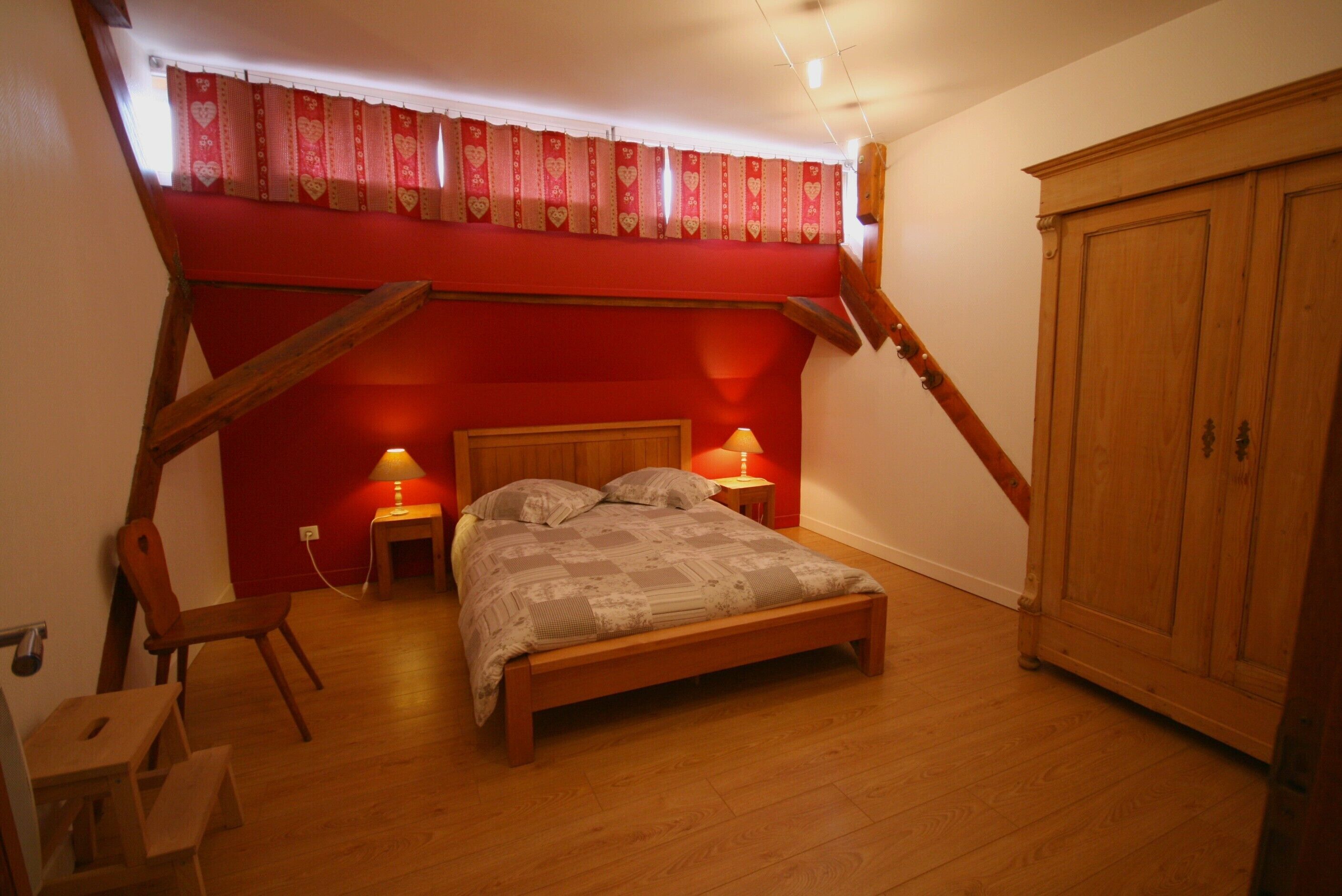 A bedroom with a wooden bed, two bedside lamps, a red accent wall, a wooden wardrobe, and a slanted ceiling.