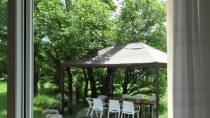 An outdoor patio with a wooden table and white chairs, a pergola, and a view of a wooded area.