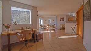 A spacious living area with a large window, a wooden desk, a dining table, and a kitchen area.