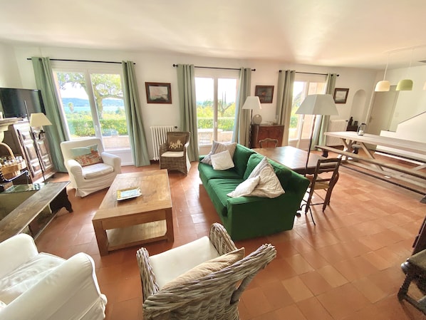 A spacious living area with a green sofa, white armchairs, a wooden coffee table, and a dining table. Large windows offer a view of the outdoors.
