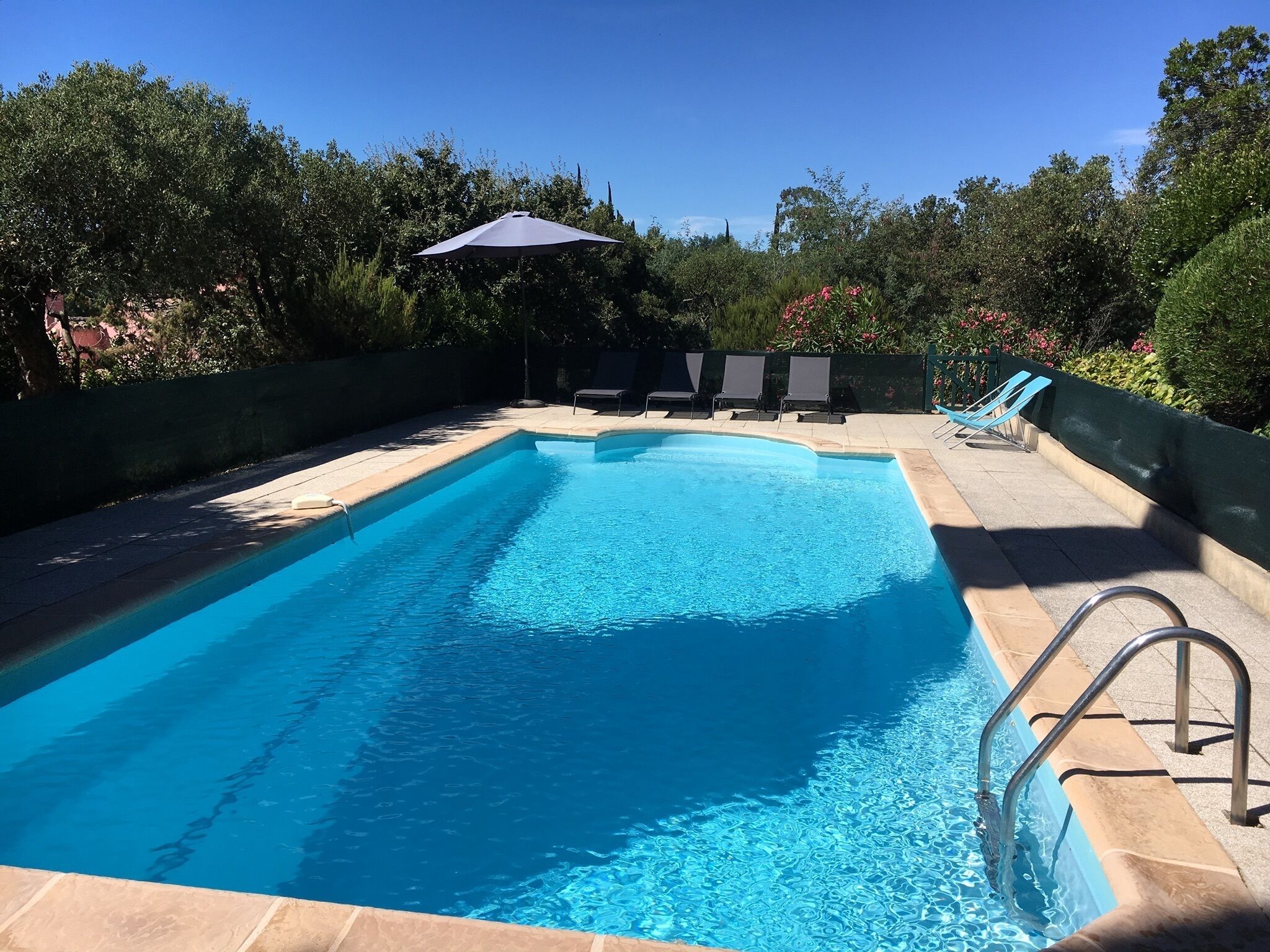 A rectangular swimming pool with clear blue water, surrounded by a tiled deck. There are lounge chairs and a covered area with an umbrella.
