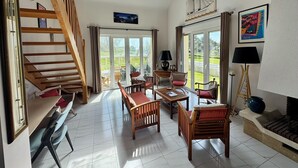 A spacious living area with a wooden staircase, a dining table, and chairs. There is a large window providing a view of the outdoors.