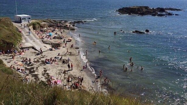 A beach with people swimming and sunbathing, rocky outcrops in the water, and a small building on the left.