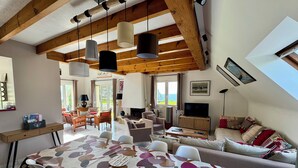 A spacious living area with a high ceiling, wooden beams, a dining table with chairs, a sofa, a television, and a fireplace.
