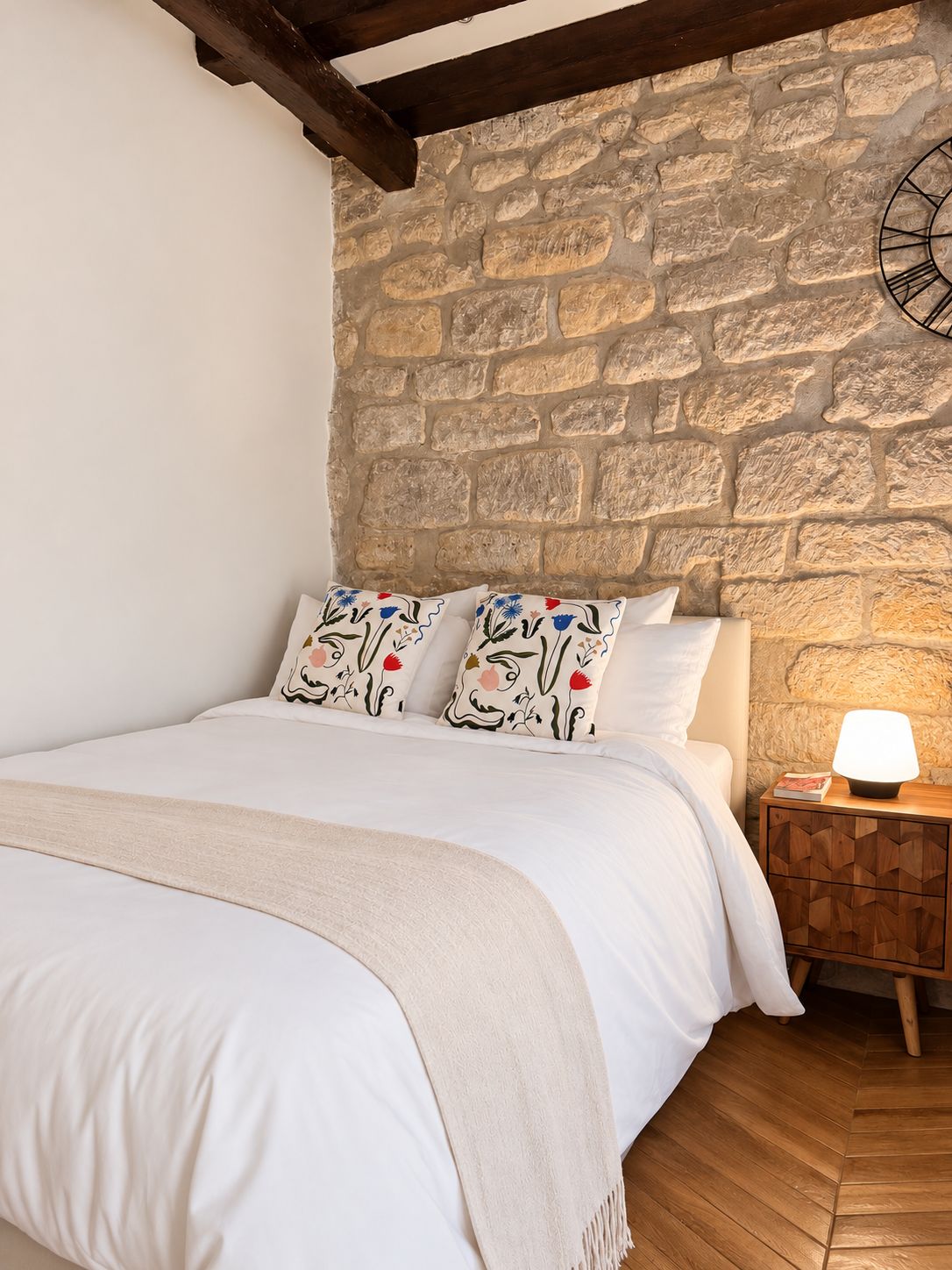 A bedroom with a stone wall, a wooden headboard, a bed with white bedding, patterned pillows, a bedside table with a lamp, and a wooden floor.