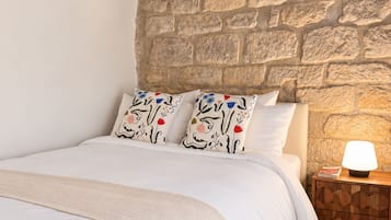 A bedroom with a stone wall, a wooden headboard, a bed with white bedding, patterned pillows, a bedside table with a lamp, and a wooden floor.