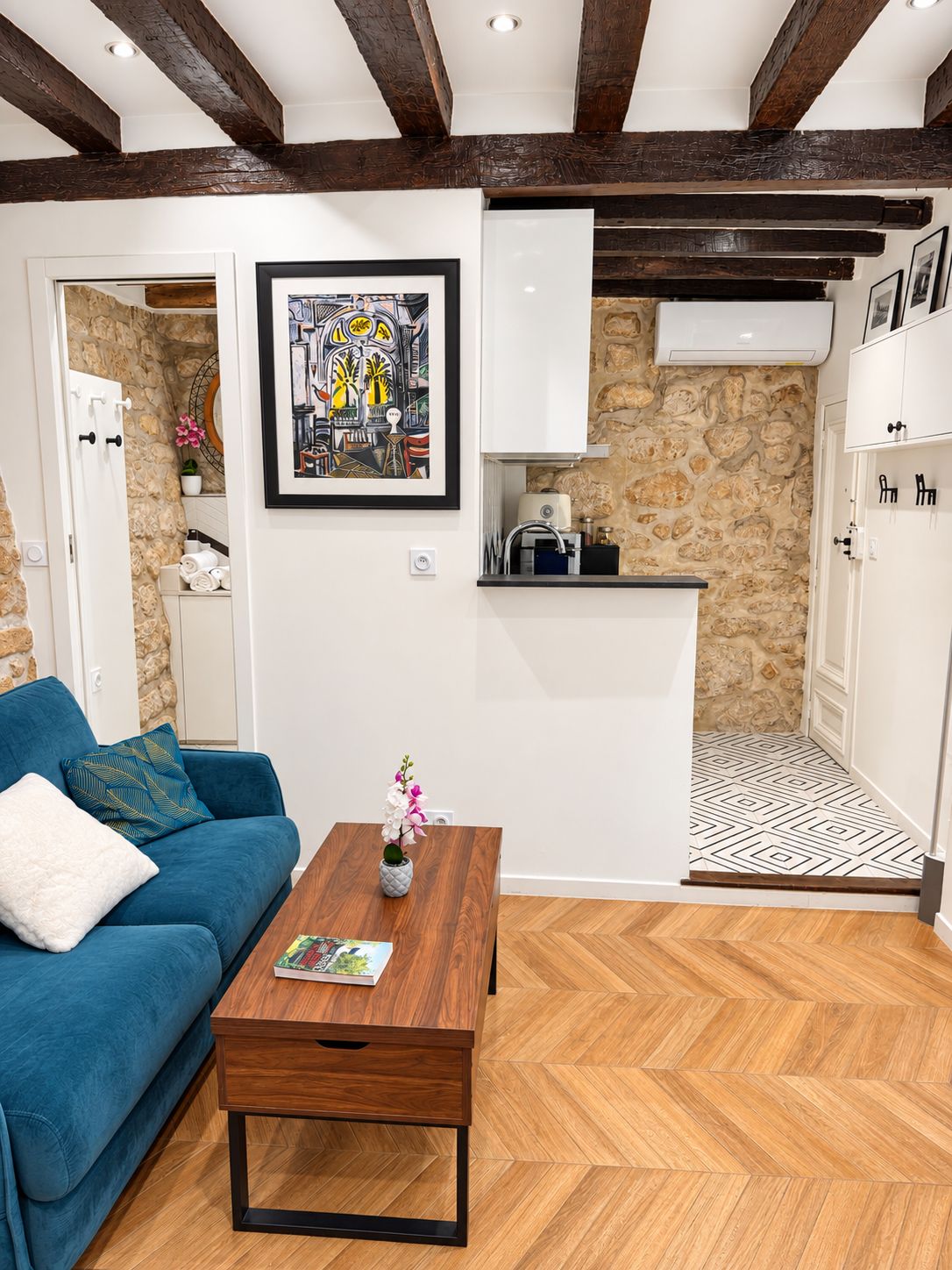 A modern living room with a blue sofa, a wooden coffee table, and a kitchen area with stone walls and wooden beams.