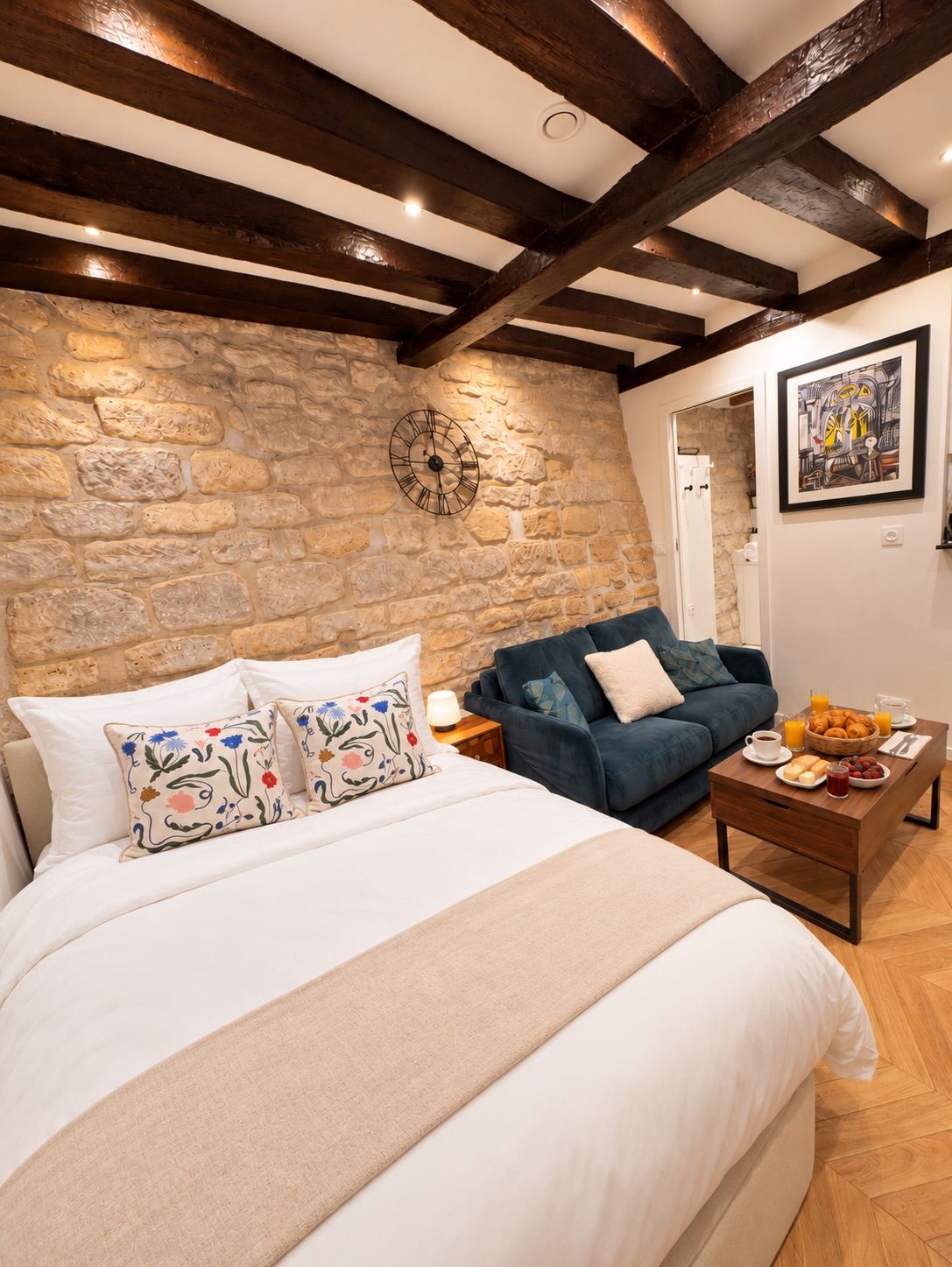 A bedroom with a stone wall, a bed with floral pillows, a blue sofa, a coffee table with breakfast items, and a clock on the wall.