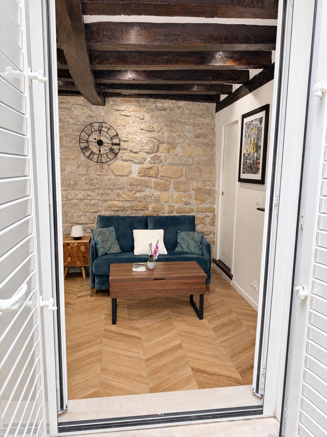 Une pièce avec un mur en pierre, un canapé bleu, une table basse en bois et une horloge au mur.