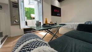 Ett modernt rum med ett glasbord, en soffa, en tv som är monterad på väggen och utsikt över utomhusmiljön genom en glasdörr.