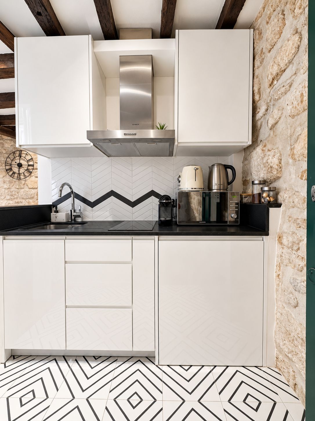 Une cuisine moderne avec des meubles blancs, une hotte en acier inoxydable, un plan de travail noir et un dosseret de carrelage disposé en motif chevrons.