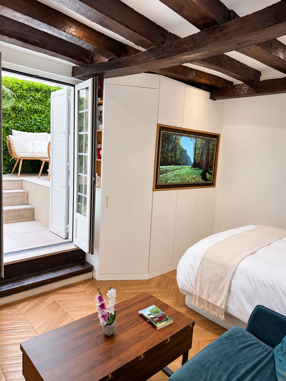 Un dormitorio con cama, una mesa de centro de madera con un libro y flores, un televisor en la pared y un puertas corredera que da a un balcón con mobiliario exterior.