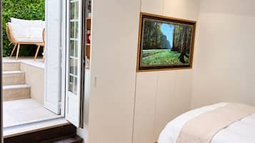 Un dormitorio con cama, una mesa de centro de madera con un libro y flores, un televisor en la pared y un puertas corredera que da a un balcón con mobiliario exterior.