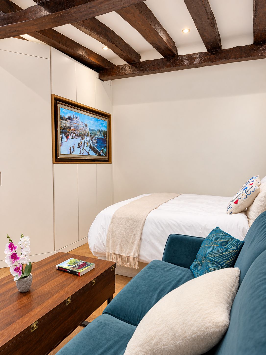 Une chambre à coucher avec un lit, une table basse en bois, un tableau encadré au mur et une fenêtre avec des poutres en bois.