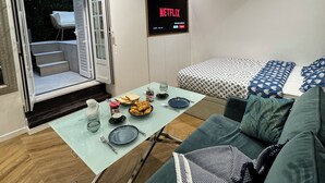 Ett modernt vardagsrum med en soffa, ett glasbord med matservis, en säng med mönstrat sänglinne, en tv som visar "NETFLIX" och en skjutdörr i glas som leder ut.