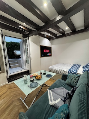 A modern living room with a sofa, a glass table with dining items, a bed with patterned bedding, a TV displaying 'NETFLIX', and a sliding glass door leading outside. - Private terrace, AC & quiet in the Latin Quarter (Paris)