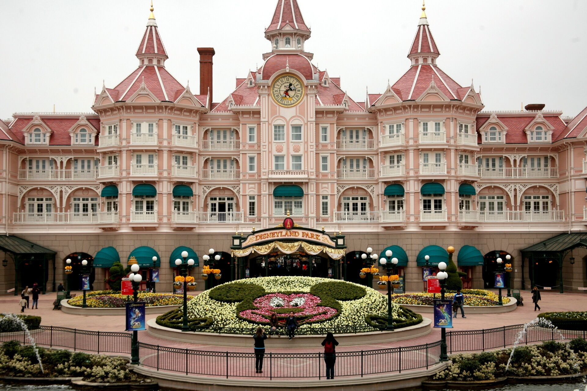 A large, pink, ornate building with a central clock tower, surrounded by a circular garden with a fountain.