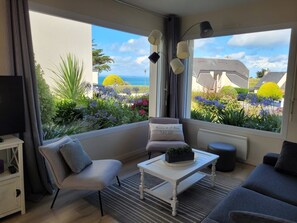 Un salon moderne comprenant un canapé, un fauteuil et une table basse, de grandes fenêtres donnant sur un jardin et des immeubles, ainsi qu’un téléviseur à écran plat posé sur un meuble.