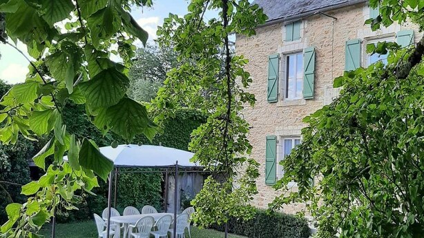 A stone building with green shutters, a covered outdoor seating area with white plastic chairs, and lush greenery. - Beautiful view of the countryside house 8 pers pool Wifi near Sarlat (Salviac)