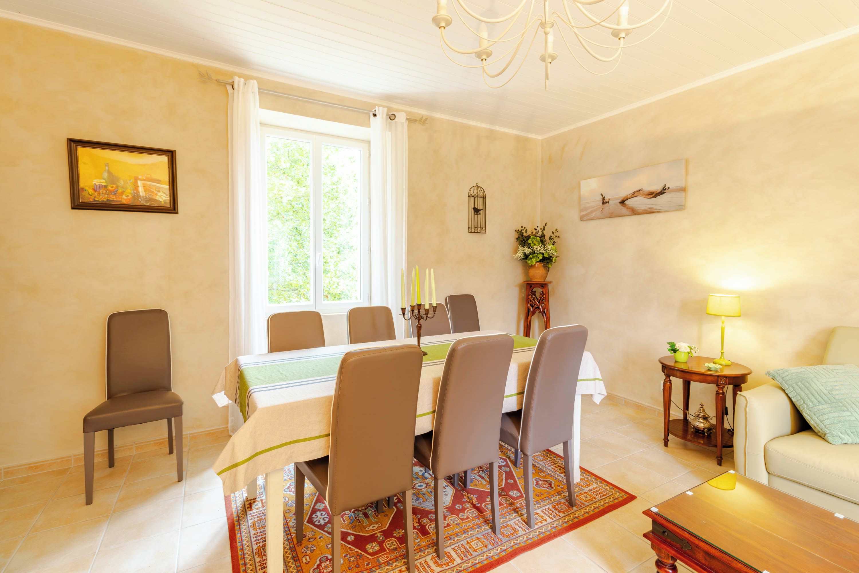 A dining area with a table set for six, surrounded by chairs, a chandelier, and a window with curtains.