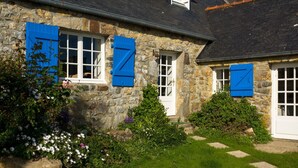 Una casa de piedra con ventanas blancas y contraventanas azules, rodeada por un jardín cuidado con diversas flores y un camino de piedra.