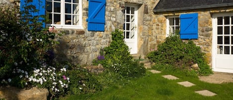 A stone house with white windows and blue shutters, surrounded by a well-maintained garden with various flowers and a stone pathway.