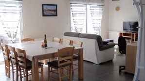 Ein Essbereich mit Tisch, Stühlen und einer Champagnerflasche. Im Wohnbereich befindet sich eine Couch und ein Fernseher.