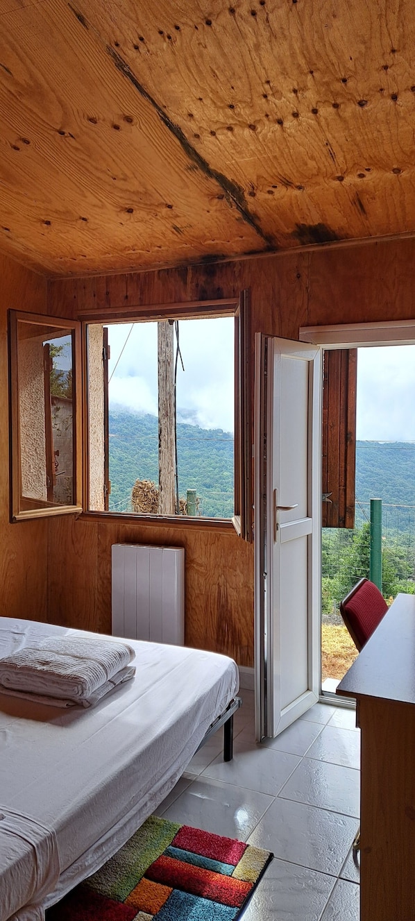 A wooden cabin room with a bed, a window with a view of mountains, and a door leading outside.