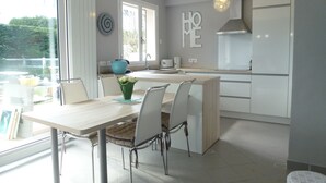 Eine moderne Küche mit weißen Schränken, einem Essbereich mit Tisch und Stühlen sowie einem Blick ins Freie durch große Fenster.