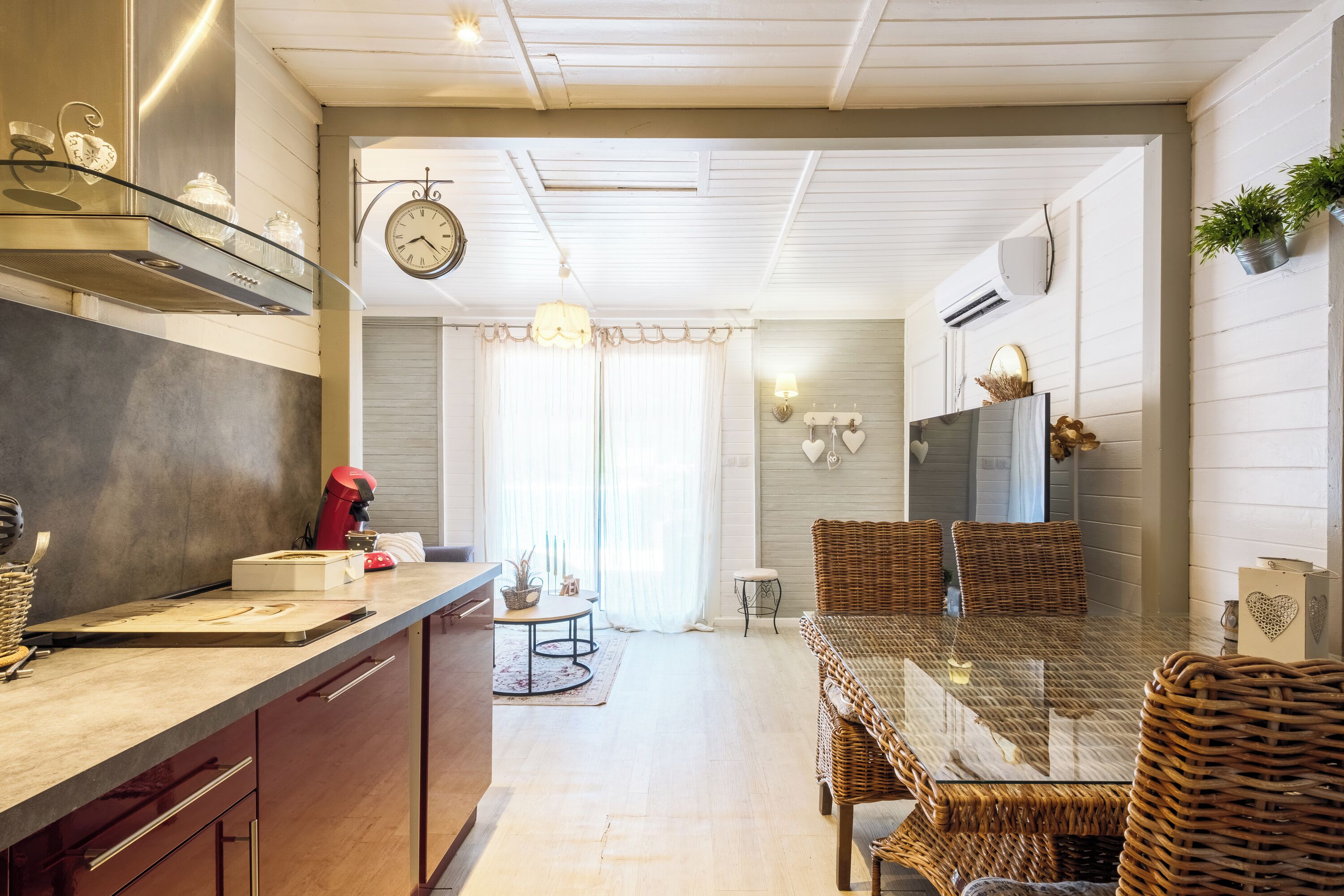 A modern kitchen with a dining area, featuring a glass-top table, wicker chairs, and a clock on the wall.